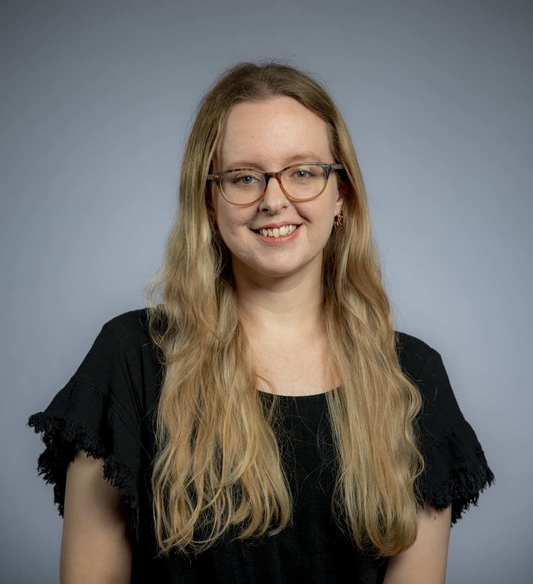 a woman with long blonde hair wearing glasses and a black top smiles against a gray background