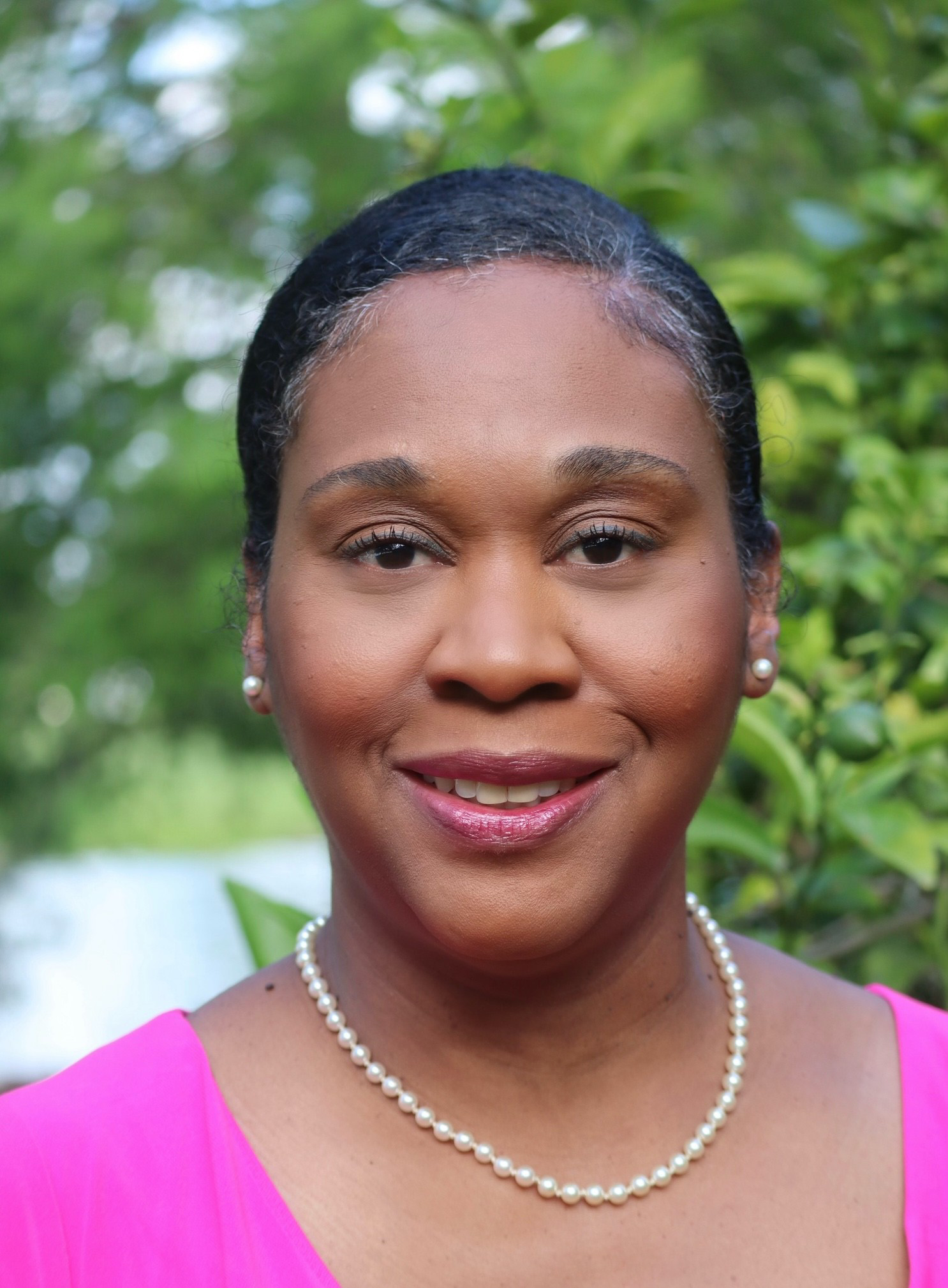 headshot of a woman smiling and wearing pearl earrings and necklace with a pink dress, standing in front of greenery outdoors